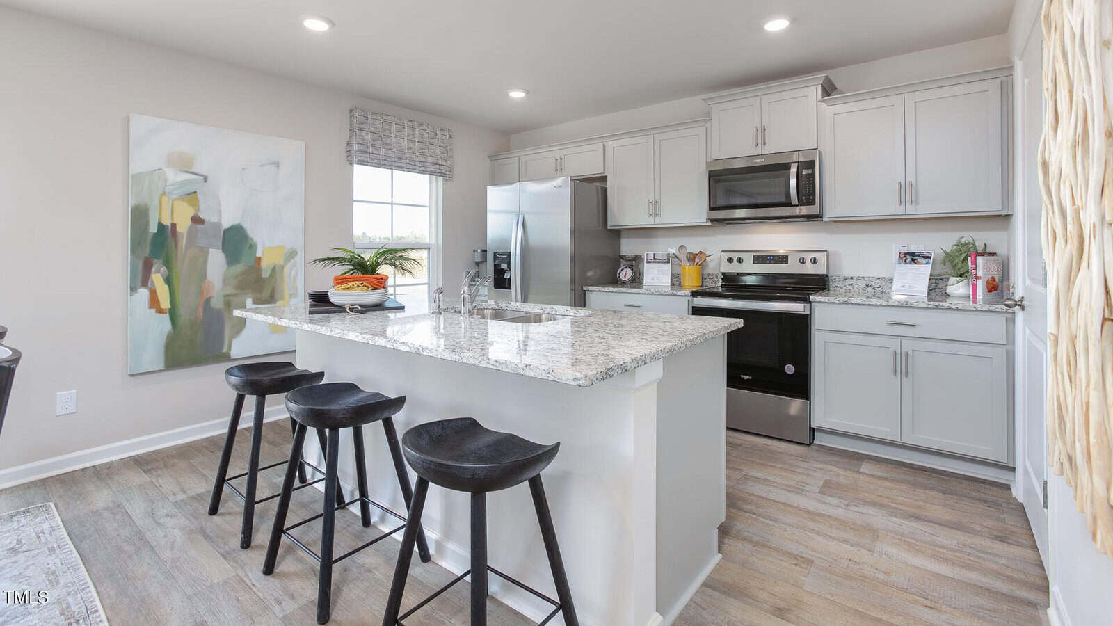Modern kitchen with light gray cabinets, stainless steel appliances, and a center island with three black stools. Abstract art hangs on the left wall, and a window brings in natural light. Light wood flooring throughout.