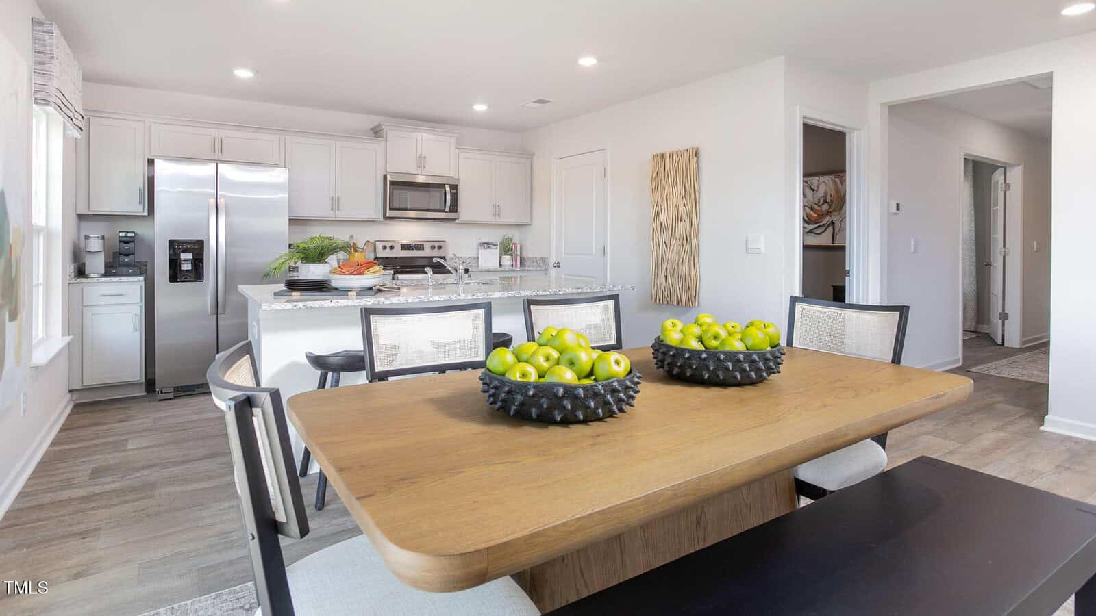 A modern kitchen and dining area with a wooden table holding two bowls of green apples, white cabinets, stainless steel appliances, and a granite countertop with barstools in the background.
