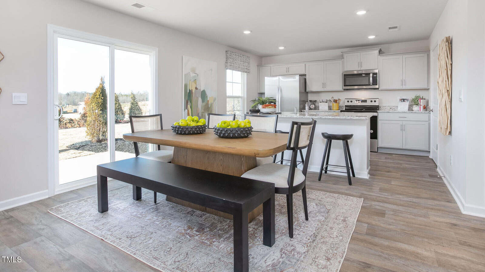 A modern kitchen and dining area features a wooden table with a bench and chairs, bowls of green apples, white cabinets, stainless steel appliances, and a sliding glass door leading outside.
