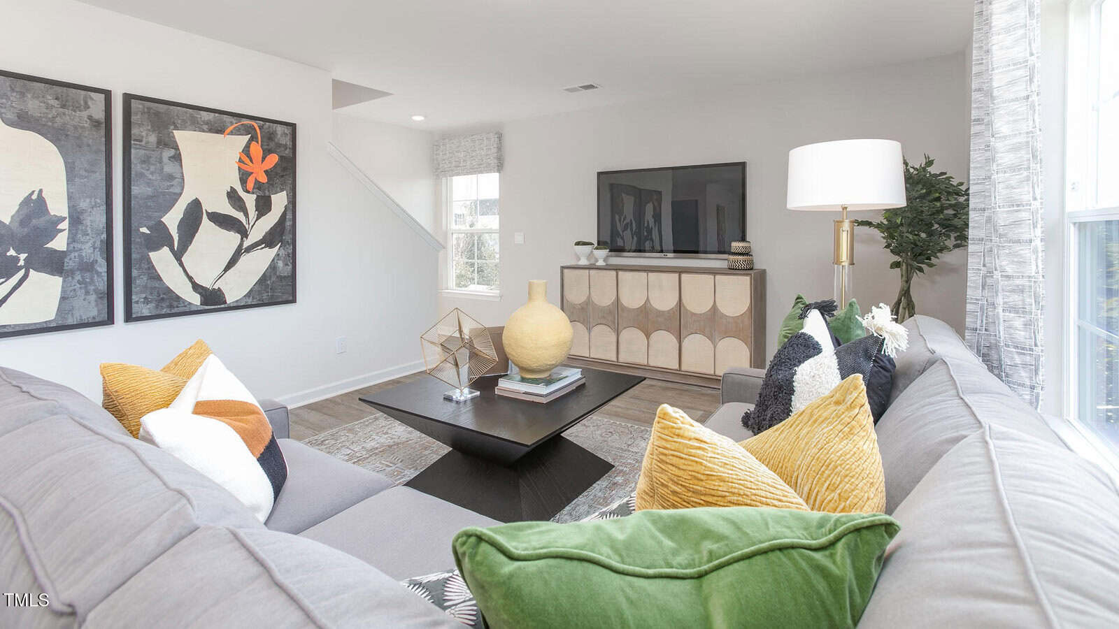 A bright living room with a gray sectional sofa decorated with colorful pillows, a black coffee table, a wooden sideboard with a TV on top, large abstract wall art, and a window letting in natural light.