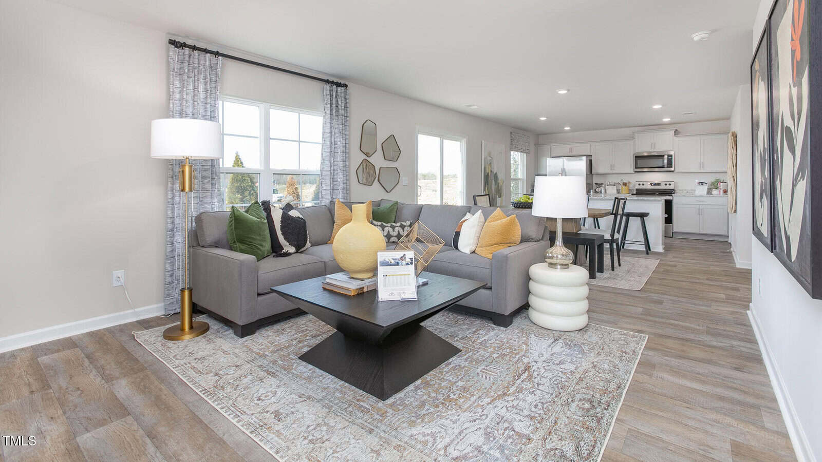 A modern open-concept living room with a gray sectional sofa, yellow and white pillows, a black coffee table, and a floor lamp, adjacent to a white kitchen and dining area with wood-style flooring.