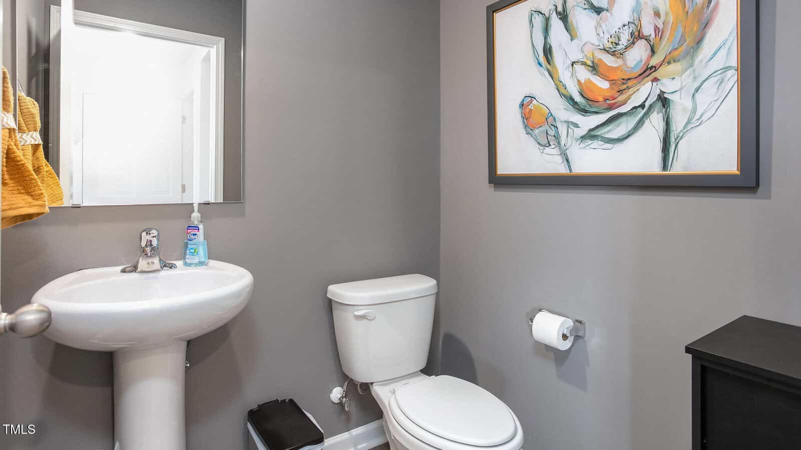 A small bathroom with gray walls, a white pedestal sink, a white toilet, a wall-mounted toilet paper holder, a soap dispenser, and a large floral painting above the toilet.