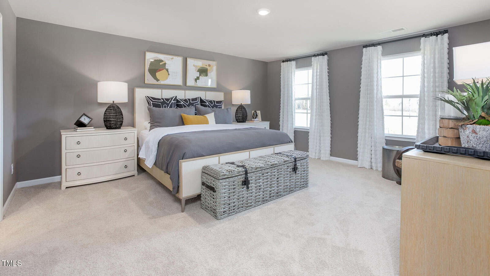 Spacious bedroom with gray walls, a large bed with gray and white bedding, two nightstands with lamps, a wicker trunk at the foot of the bed, and two large windows with white curtains.