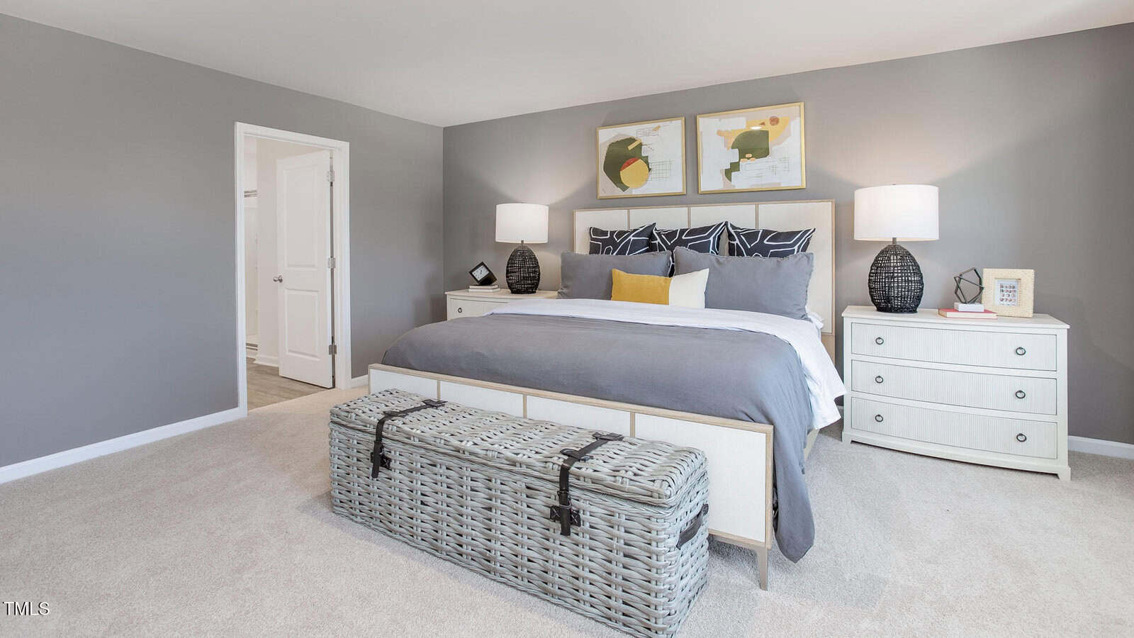 Modern bedroom with a large bed, gray bedding, two white nightstands with lamps, wicker storage bench at the foot of the bed, and abstract art above the headboard. Walls are light gray and carpet is beige.