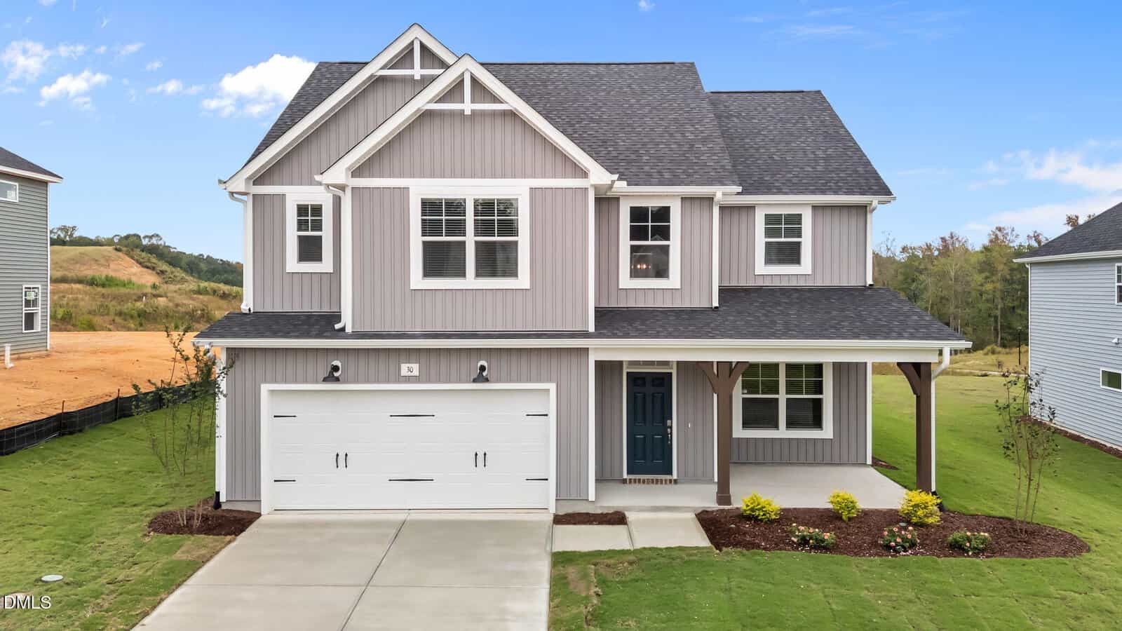 Two-story gray house with white trim, a covered front porch, blue front door, and a two-car garage. Neatly landscaped yard with shrubs and grass, set against a clear blue sky and neighboring houses.