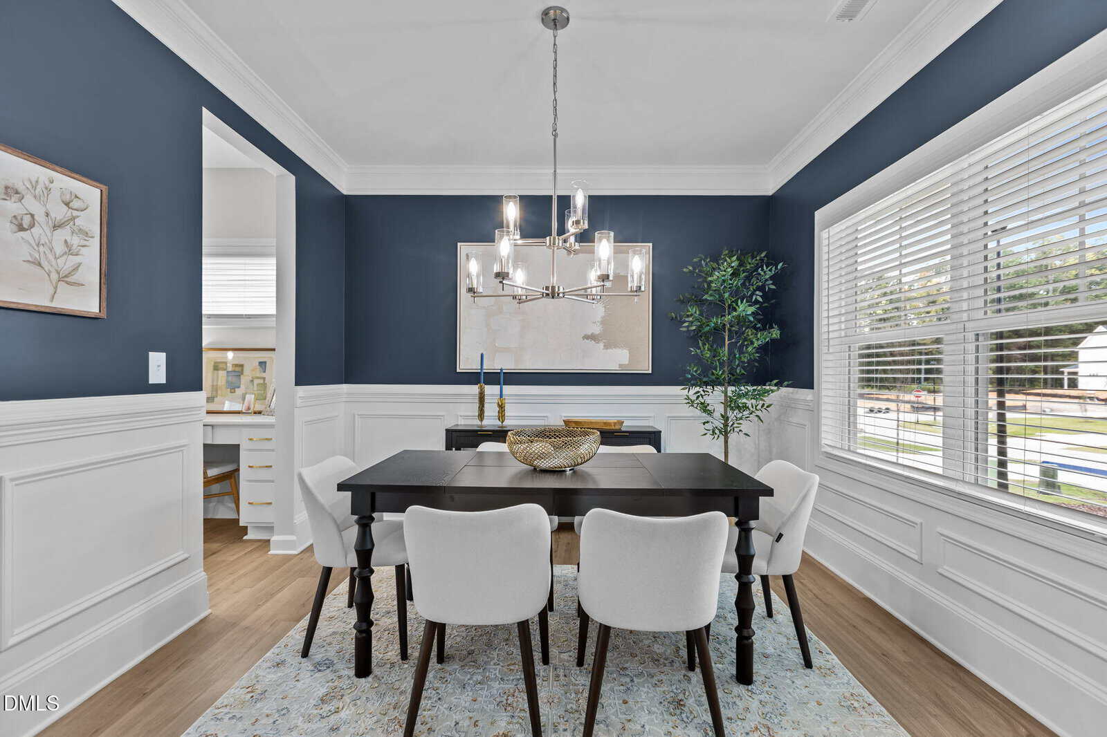 A modern dining room with navy blue walls, white wainscoting, a black dining table, six white chairs, a rug, a chandelier, wall art, a potted plant, and large windows letting in natural light.