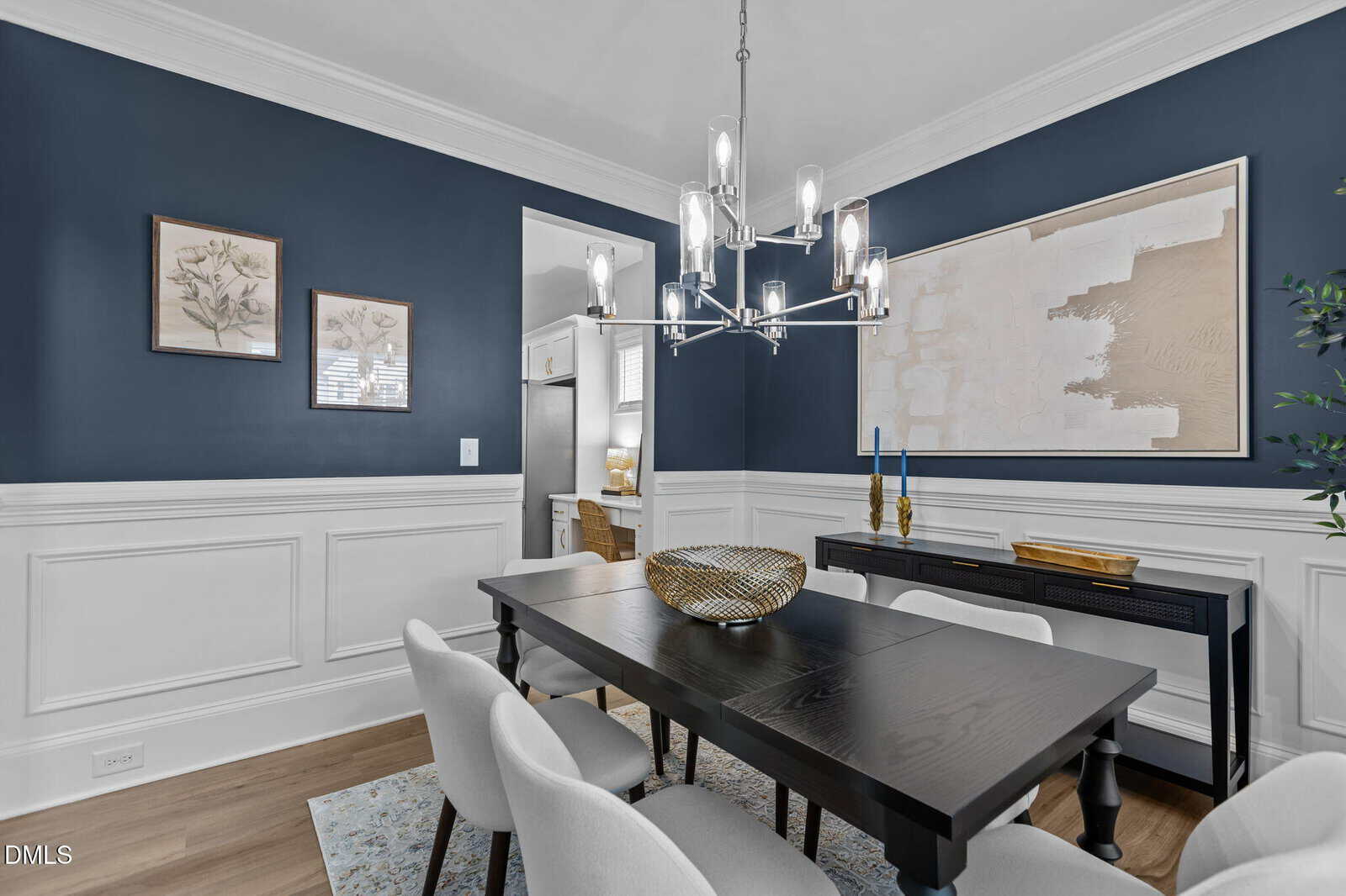 A modern dining room with navy blue and white walls, a black table with six white chairs, contemporary chandelier, decorative art on the walls, and a sideboard with candles and a gold bowl.