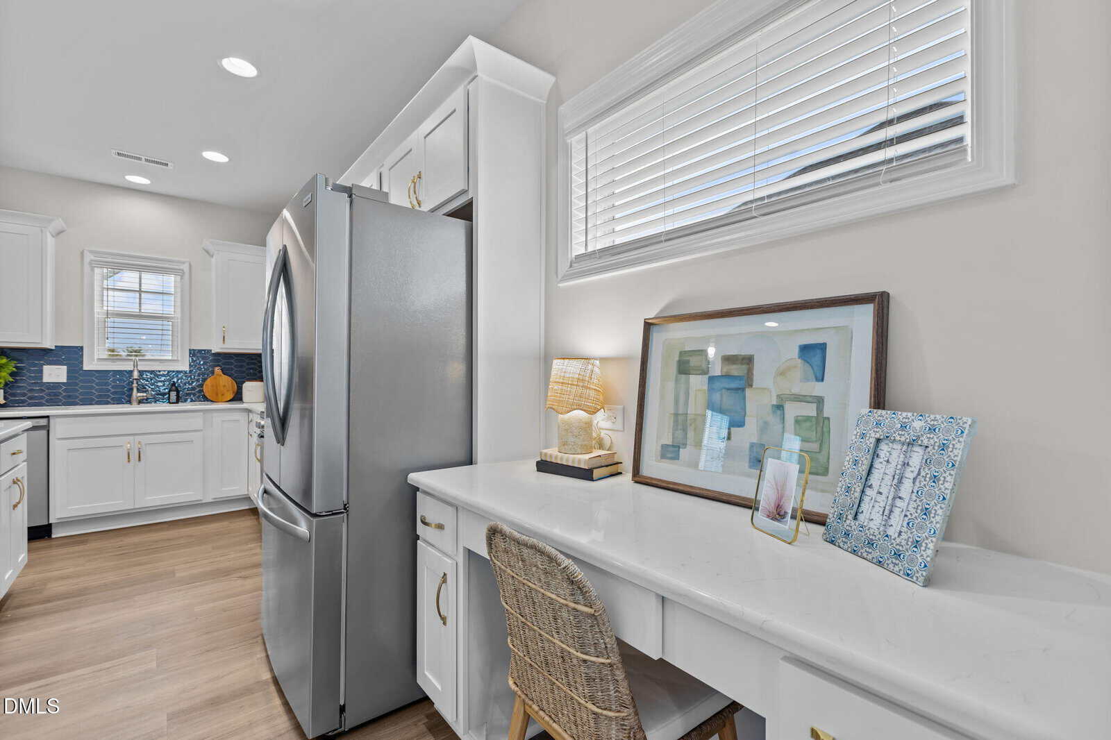A bright kitchen with white cabinets and a stainless steel fridge. A built-in desk area with a wicker chair, lamp, framed artwork, and a tablet sits under a window with blinds. Light wood floors throughout.