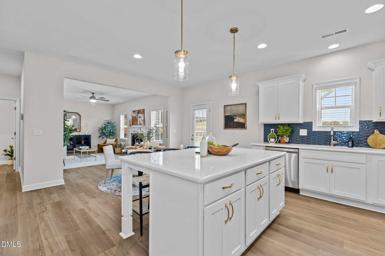 Bright modern kitchen with white cabinets, gold handles, a large island with stools, pendant lights, wood flooring, and an open view into a cozy living area with a sofa and plants.