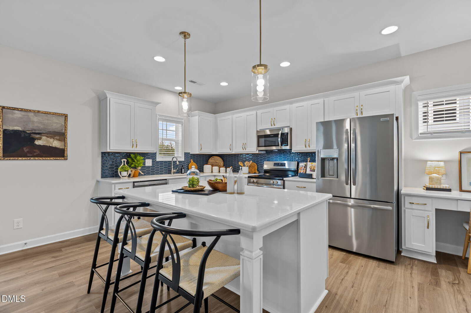 Modern kitchen with white cabinets, stainless steel appliances, an island with three black barstools, pendant lighting, wood floors, and blue tile backsplash. Decor includes plants, art, and countertop accessories.