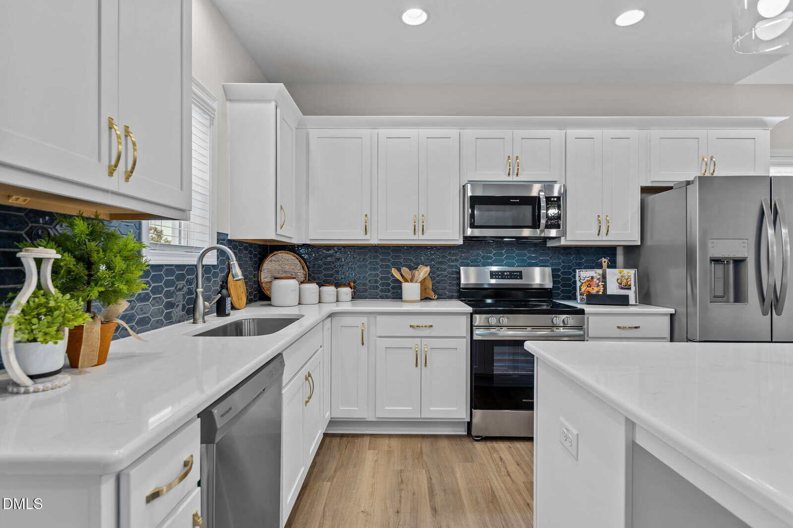 Modern kitchen with white cabinets, stainless steel appliances, blue hexagon tile backsplash, and light wood floors. Countertops are white with plants, utensils, and jars neatly arranged.