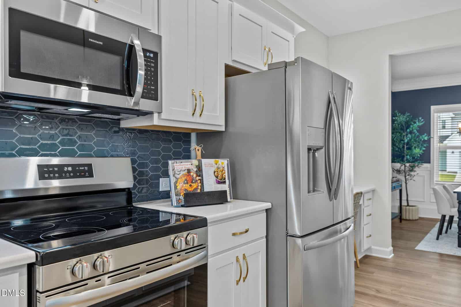 Modern kitchen with stainless steel appliances, including a stove, microwave, and refrigerator. White cabinets with gold handles, hexagonal blue tile backsplash, and a cookbook stand on the counter. Dining area visible in background.