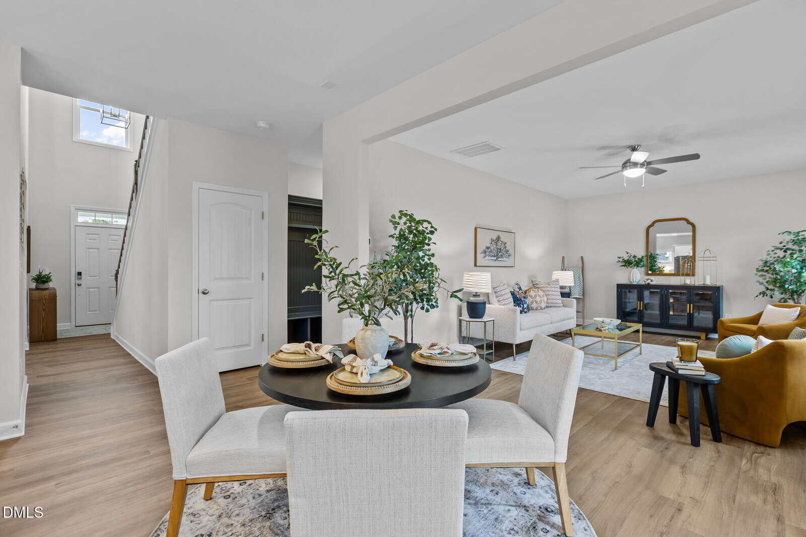 Open-concept living space with a dining area in the foreground, set for four. Beyond is a bright living room with a sofa, armchair, plants, and a ceiling fan. Wood floors and neutral decor throughout.