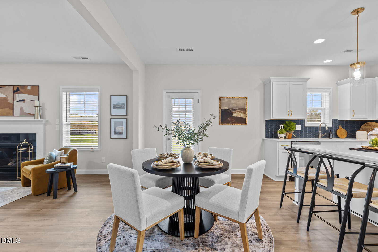 Modern open-concept kitchen and dining area with a round table set for four, white chairs, sleek kitchen island with stools, and a cozy living space with a fireplace and armchair in the background.