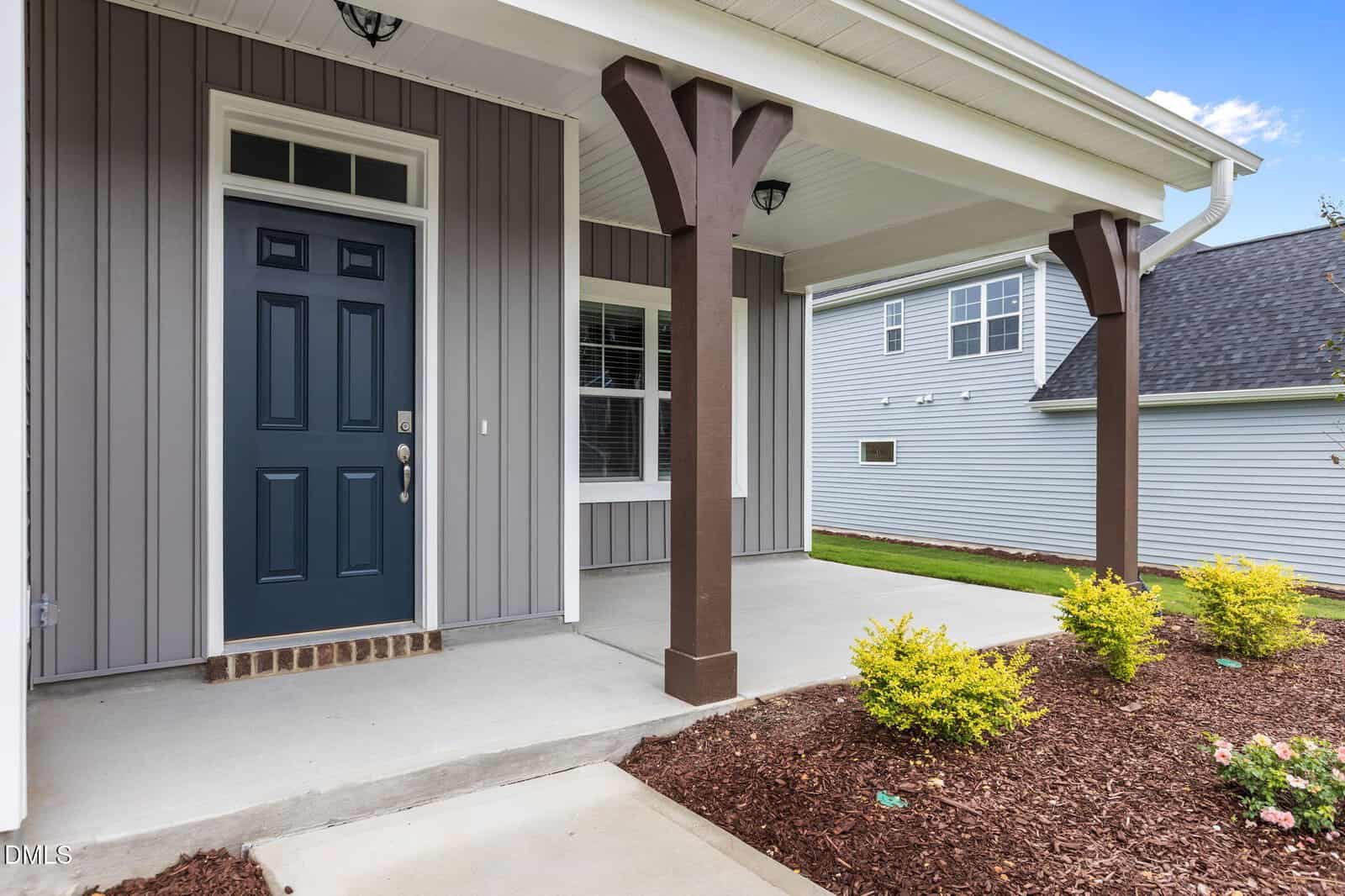 Covered front porch of a modern house with gray vertical siding, dark blue door, white trim, and mulch landscaping with small yellow-green bushes. Neighboring house is visible in the background.