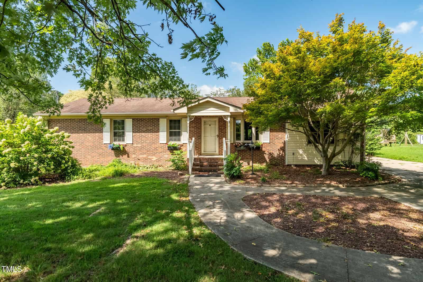 Charming single-story brick house in Henderson at 1006 Abbott Road, featuring white trim, a front porch with steps, and a brown shingle roof. A curved concrete walkway winds through lush lawn, trees, and landscaping to the entrance.