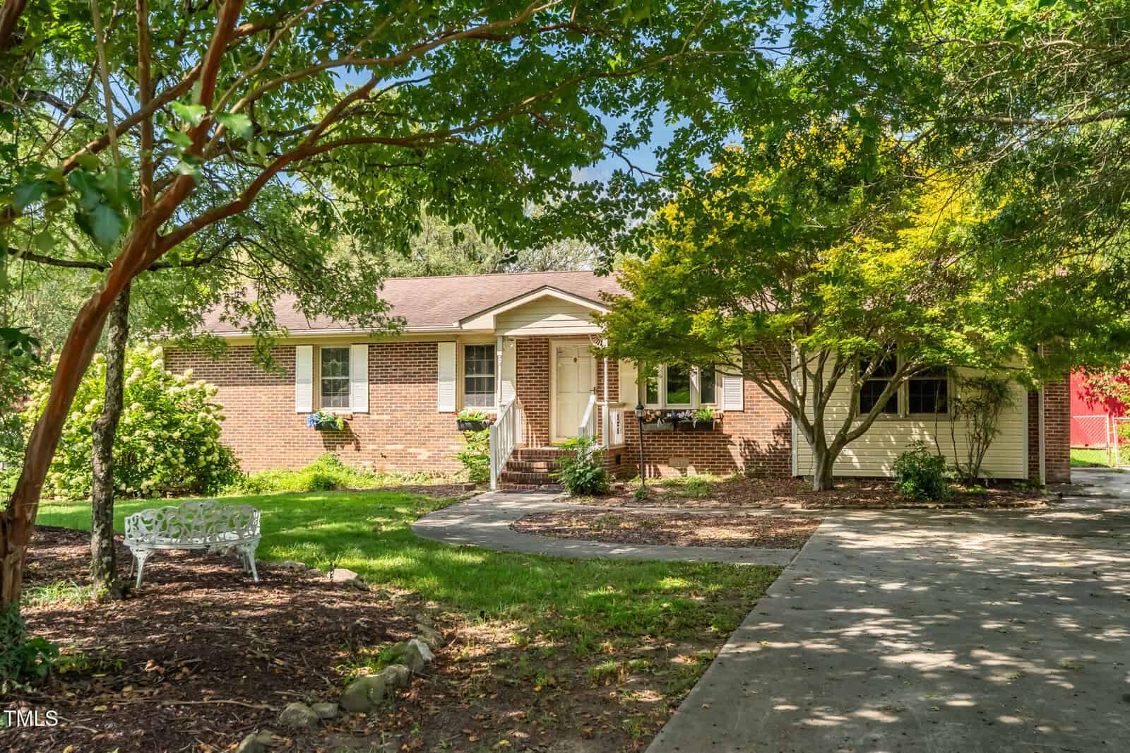 A single-story brick and siding house with a covered front porch, surrounded by green trees and landscaping. A concrete driveway and a white metal bench are visible in the foreground.