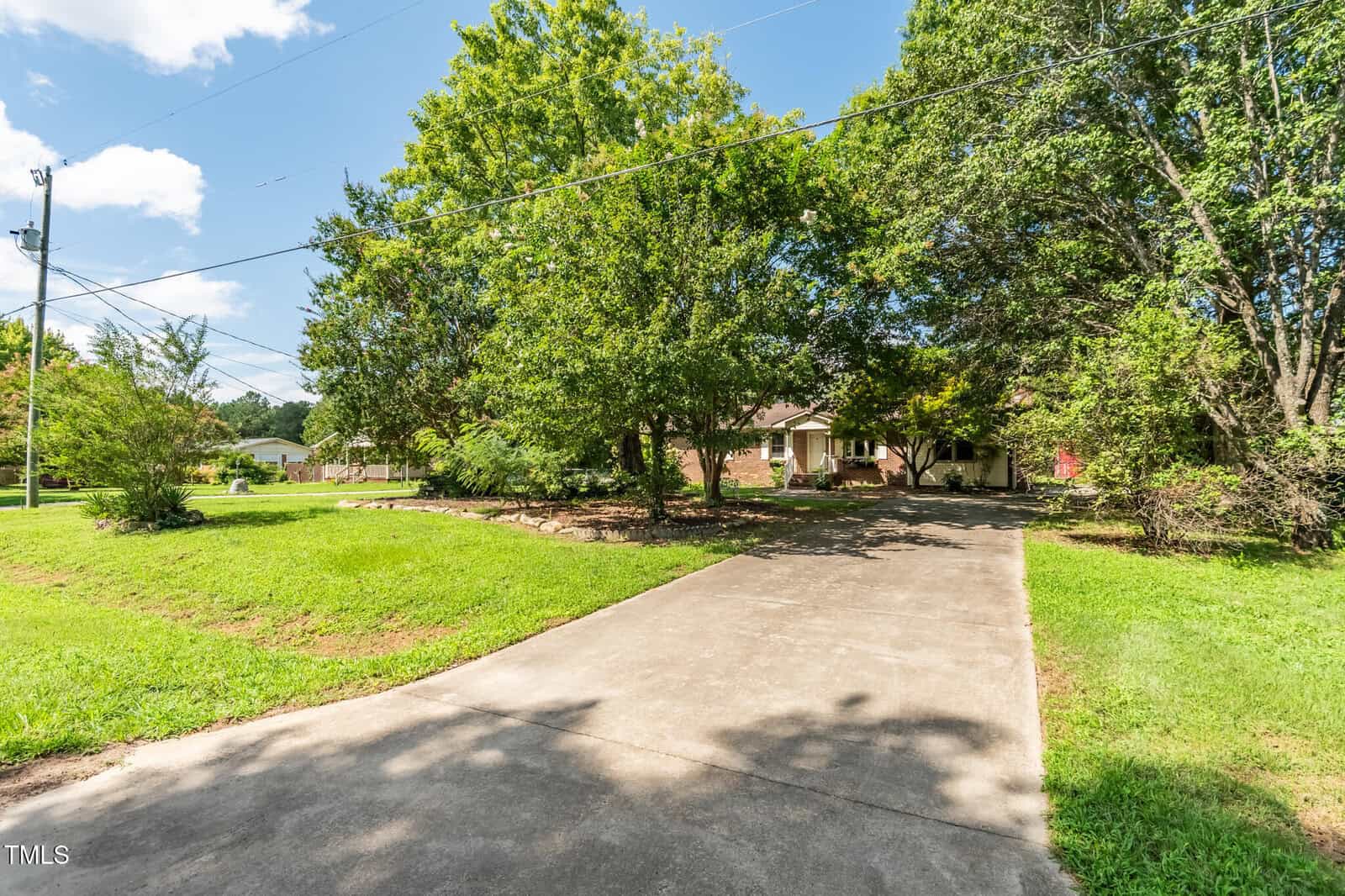 A concrete driveway leads to a single-story brick house partially obscured by large green trees, with a well-maintained lawn on either side and a clear blue sky overhead.