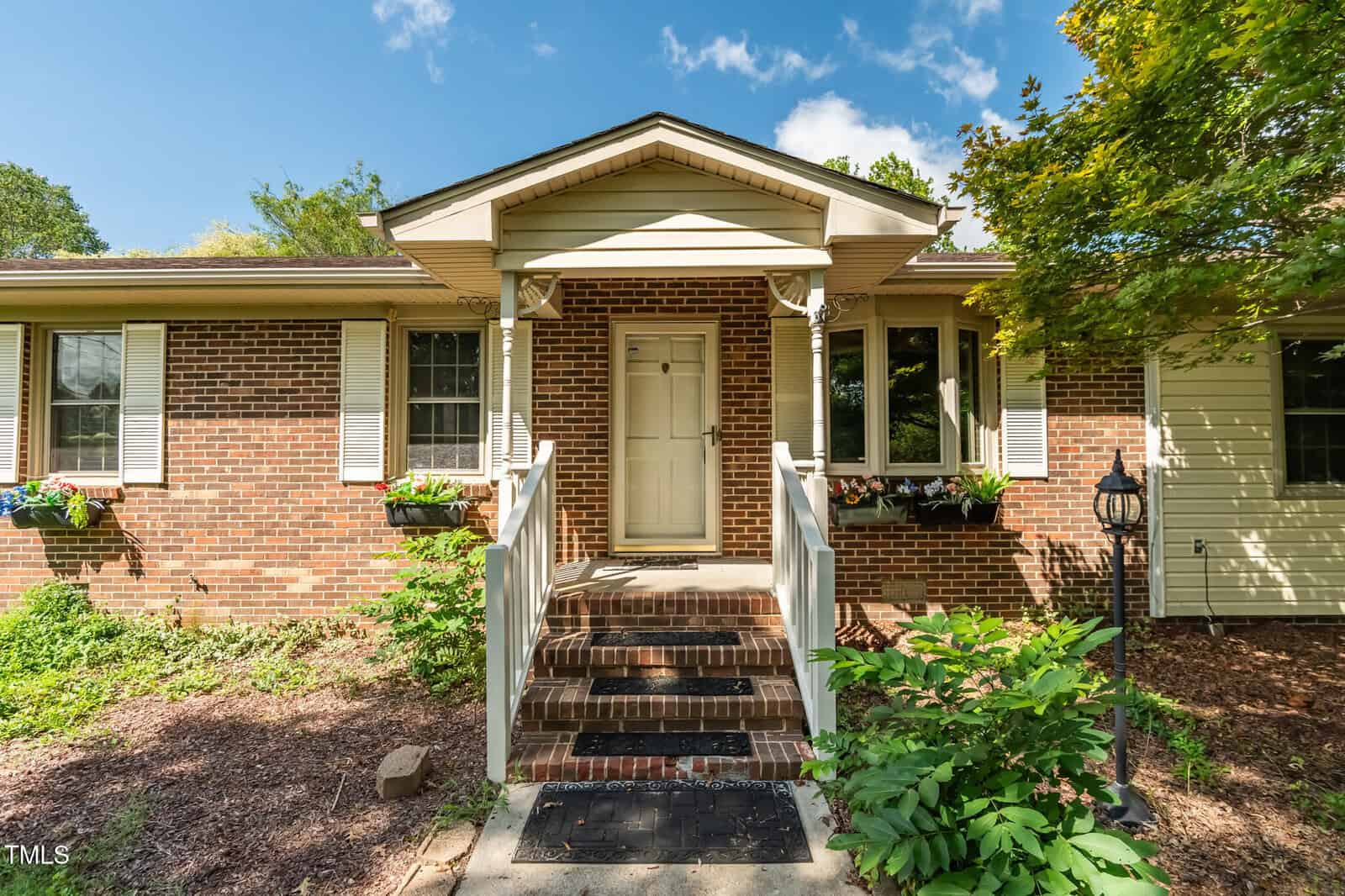 A brick house with white trim and shutters, a covered front porch, white railing, steps with black doormat, flower boxes under windows, and a lamp post on the right, surrounded by landscaping and greenery.