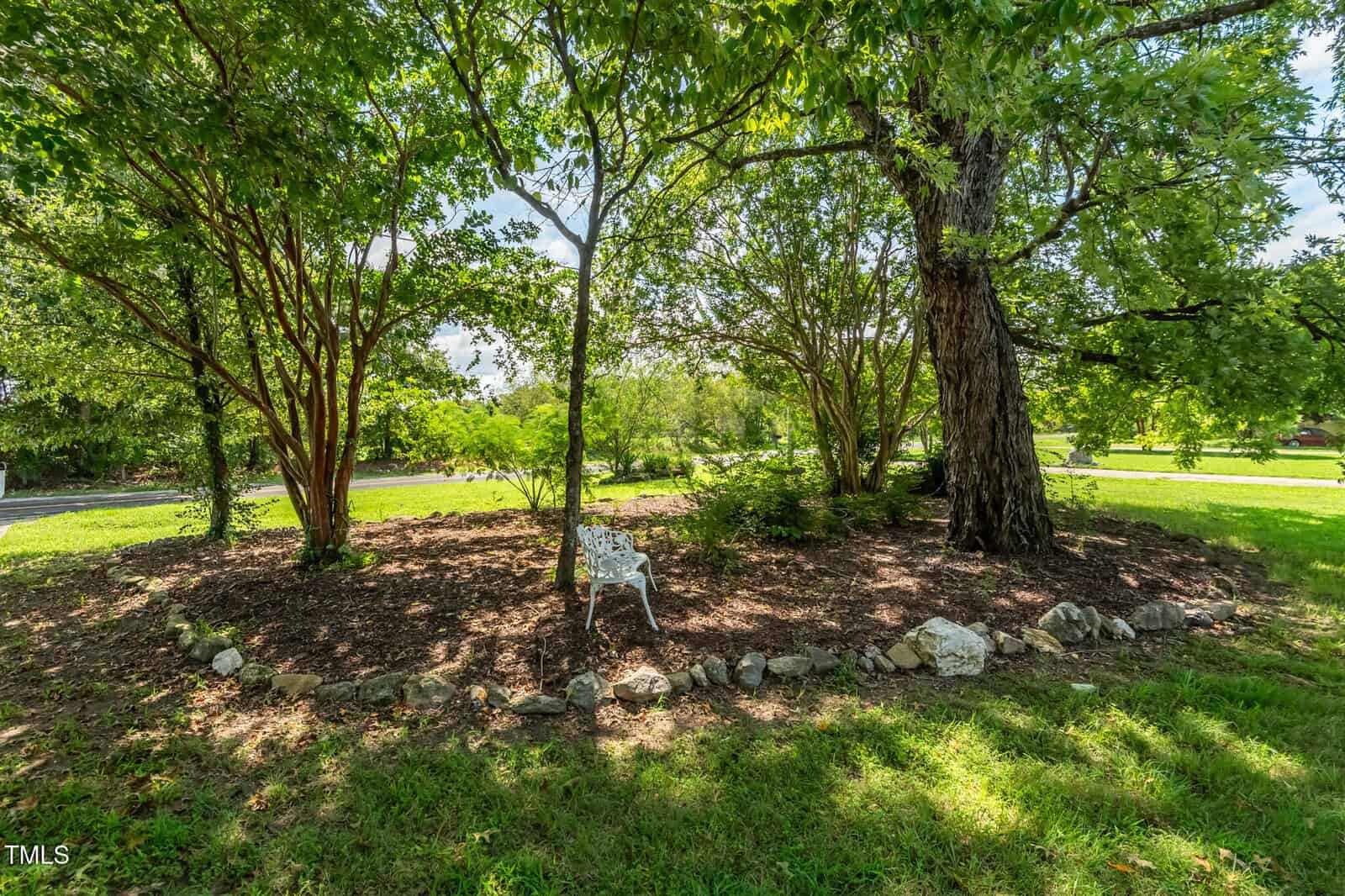 A white metal chair sits on a mulched area under large leafy trees, surrounded by a border of rocks, with green grass and a road visible in the background.