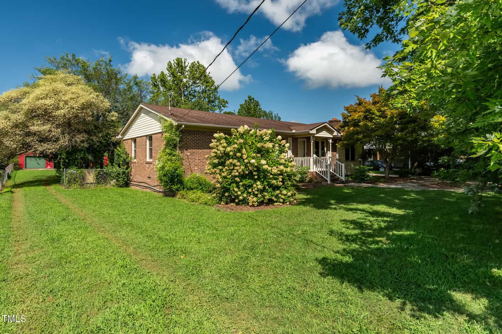A single-story brick house with a covered front porch, surrounded by green grass, shrubs, and trees under a blue sky with scattered clouds. A driveway and a red shed are visible on the left side.