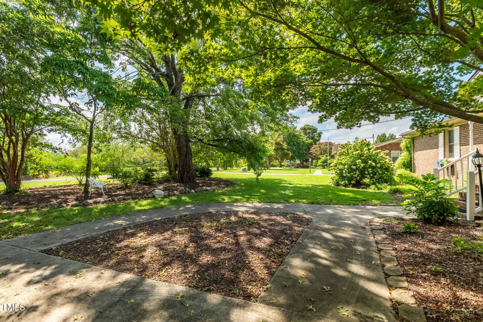 A shaded patio area with concrete paths and mulch beds is surrounded by green grass, large trees, and a brick house on the right side under a sunny sky.