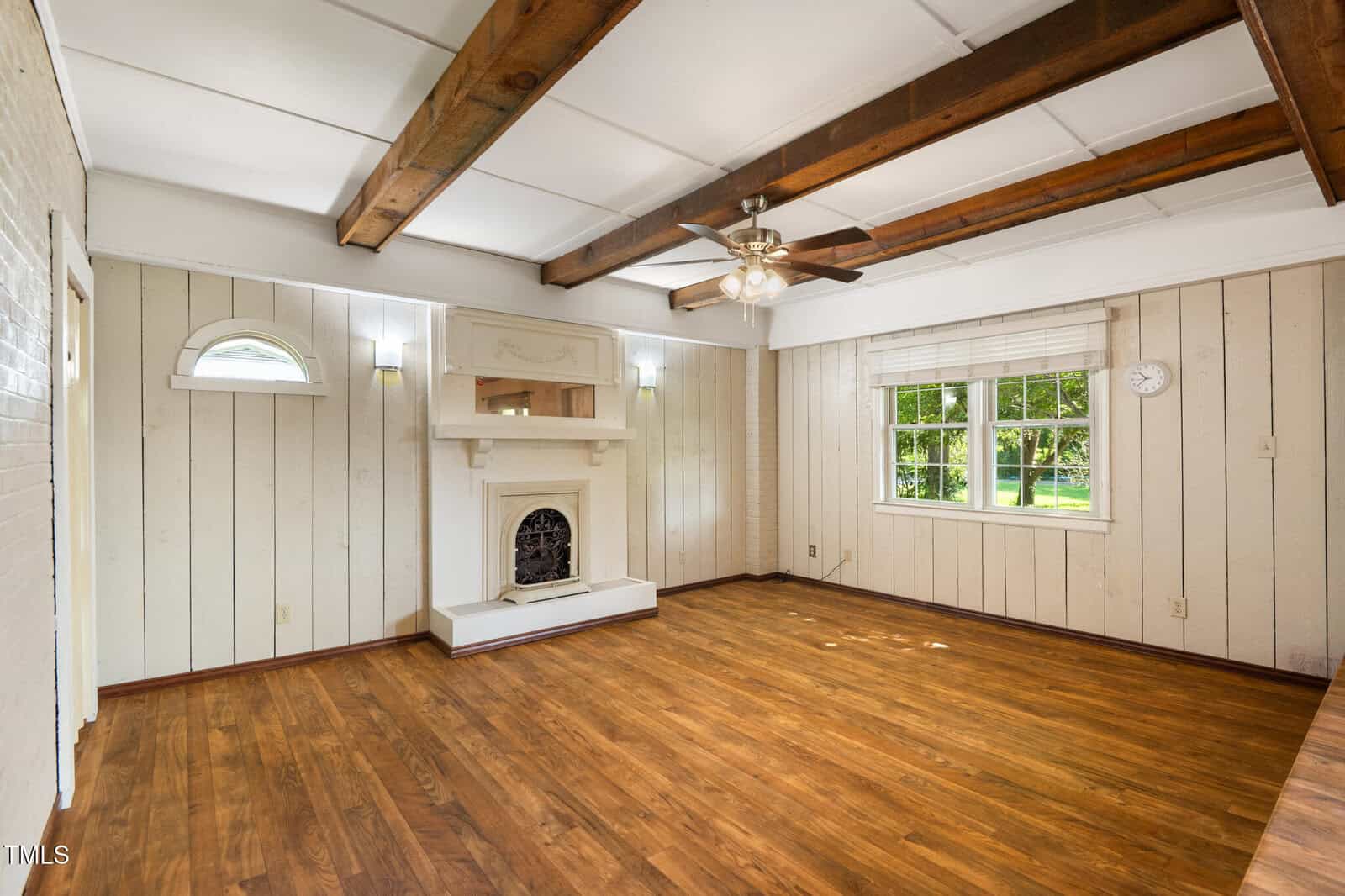 Bright living room with wood floors, white paneled walls, wood ceiling beams, a ceiling fan, fireplace with shelf, and large window with white blinds. Small wall clock and greenery visible outside the window.