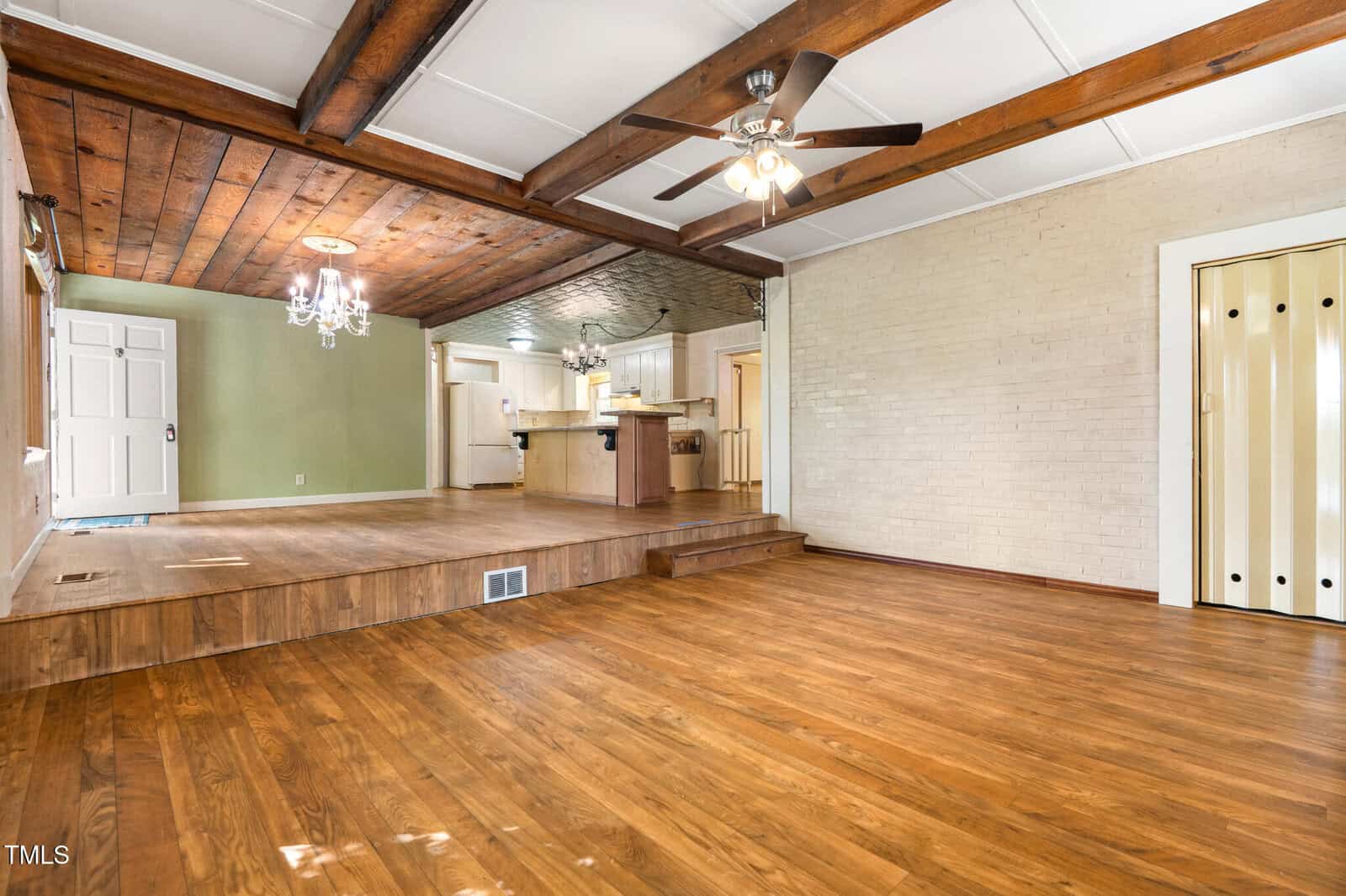 Spacious room with wood floors, exposed wood ceiling beams, ceiling fan, two chandeliers, white brick wall, and an elevated area leading to an open kitchen with bar seating.