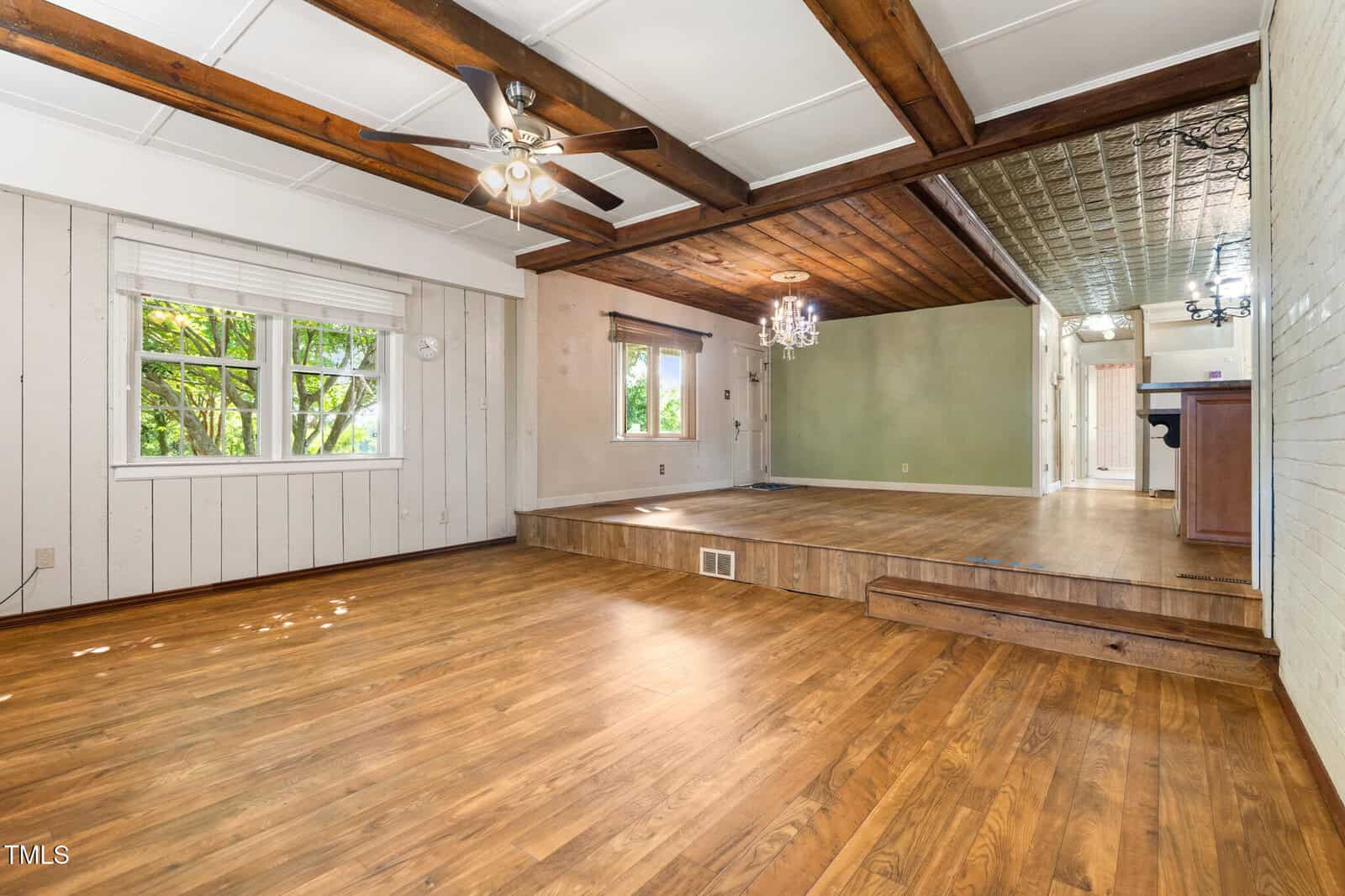 Spacious room with wood floors, coffered ceiling, ceiling fan, raised dining area, green accent wall, and large windows letting in natural light.