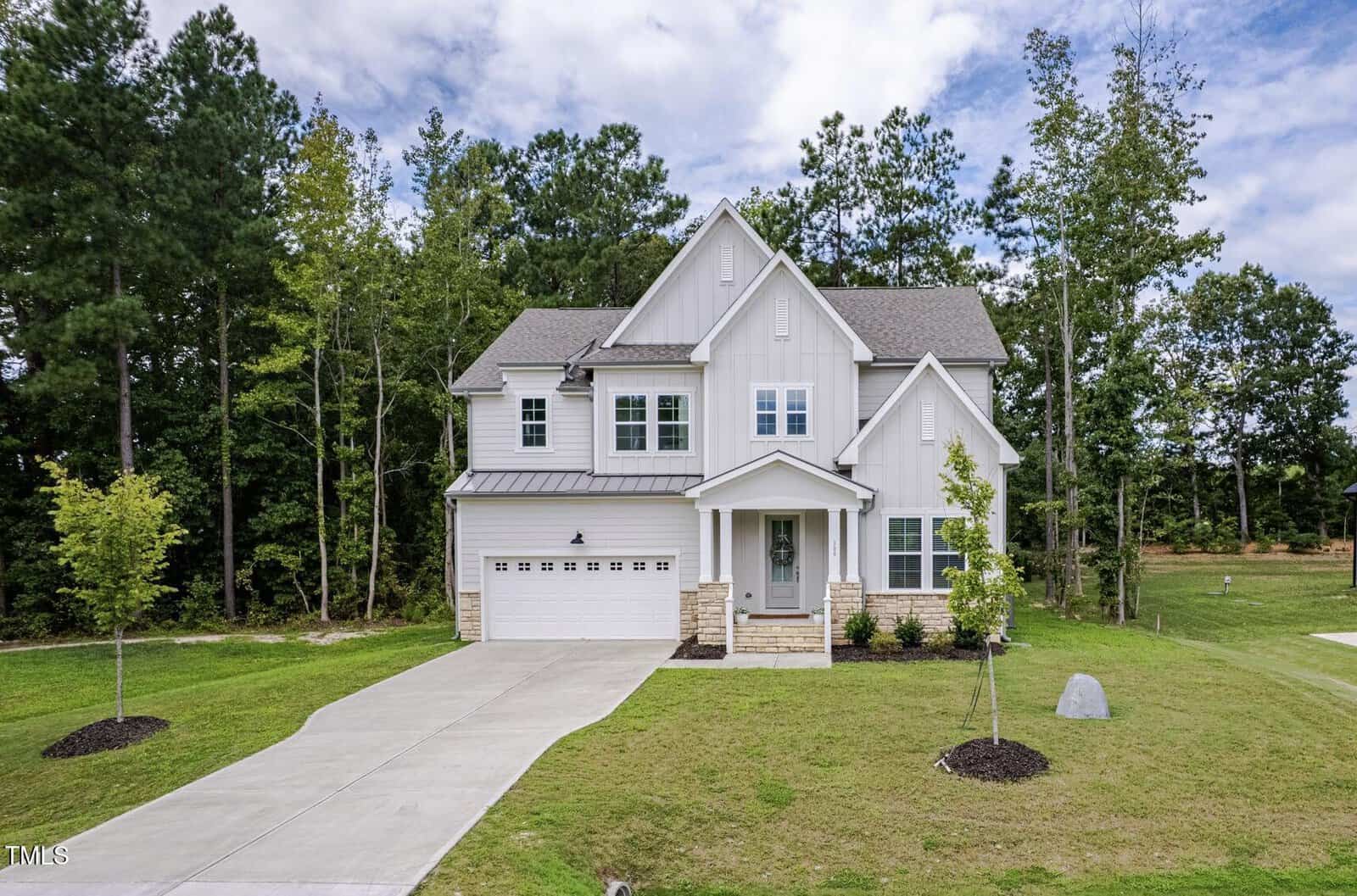 Two-story white house with a front porch, double garage, and gray roof. The house sits on a green lawn with young trees and a concrete driveway, bordered by woods and partly cloudy sky in the background.