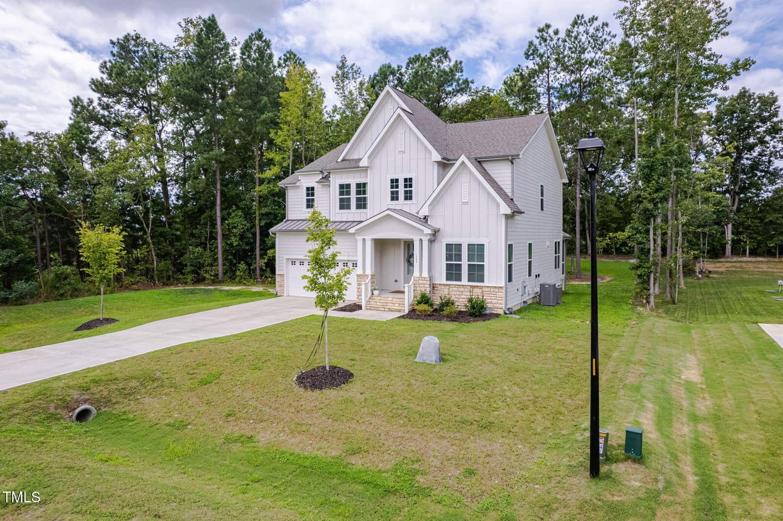 Two-story white house with a gray roof, attached garage, and front porch, set on a large grassy lot with a few young trees. Surrounded by woods and a street lamp near the driveway. Sky is partly cloudy.
