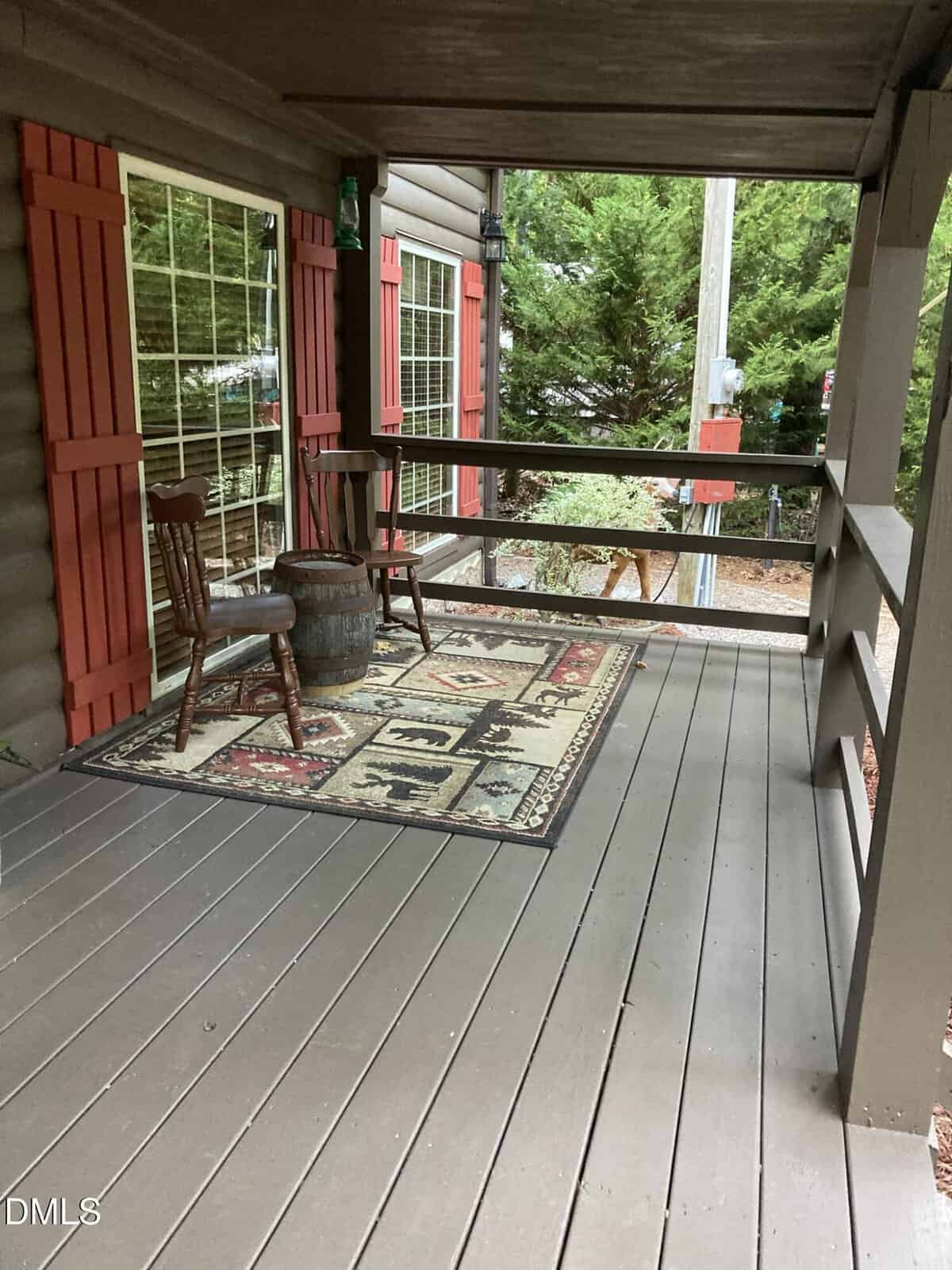 A wooden cabin porch with a rug, two wooden chairs, and a barrel table. The porch has railings, red window shutters, and overlooks greenery and a utility pole in the background.