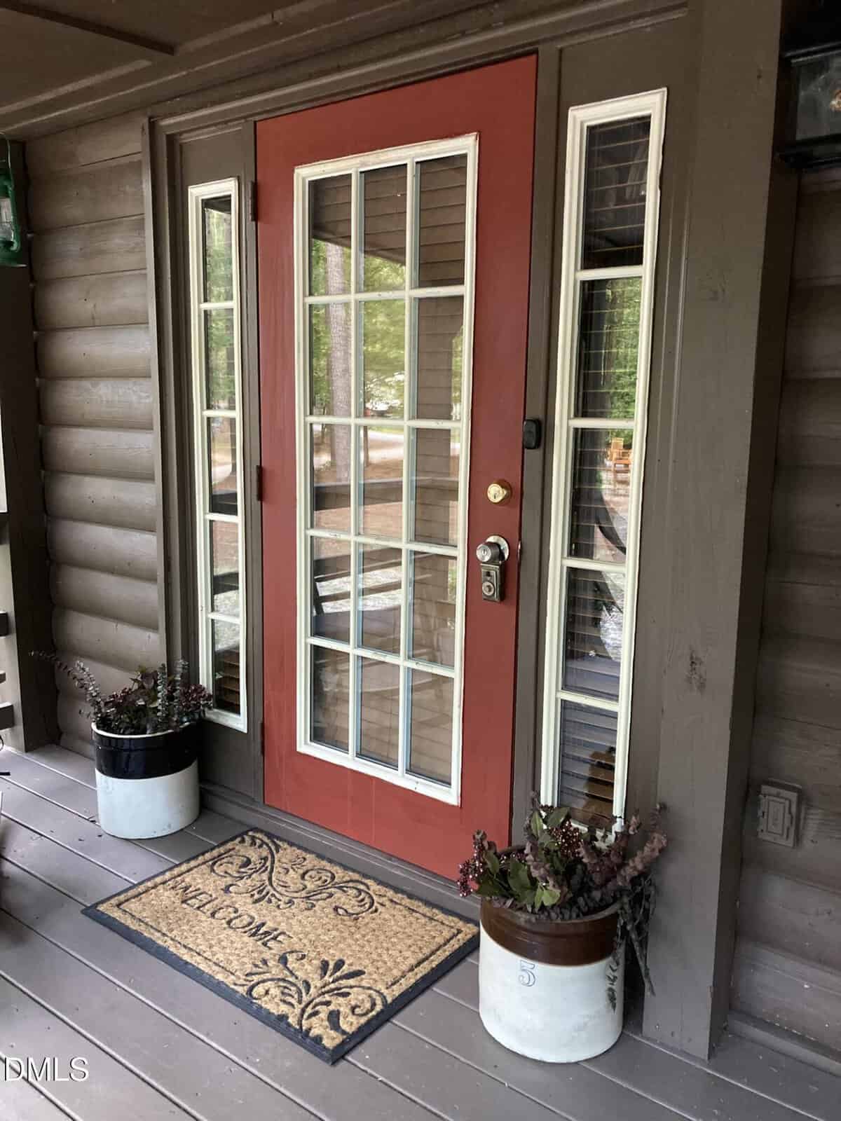 A red front door with glass panels and white trim is flanked by two tall narrow windows. Two potted plants and a "Welcome" doormat sit on the brown wooden porch in front of the door.