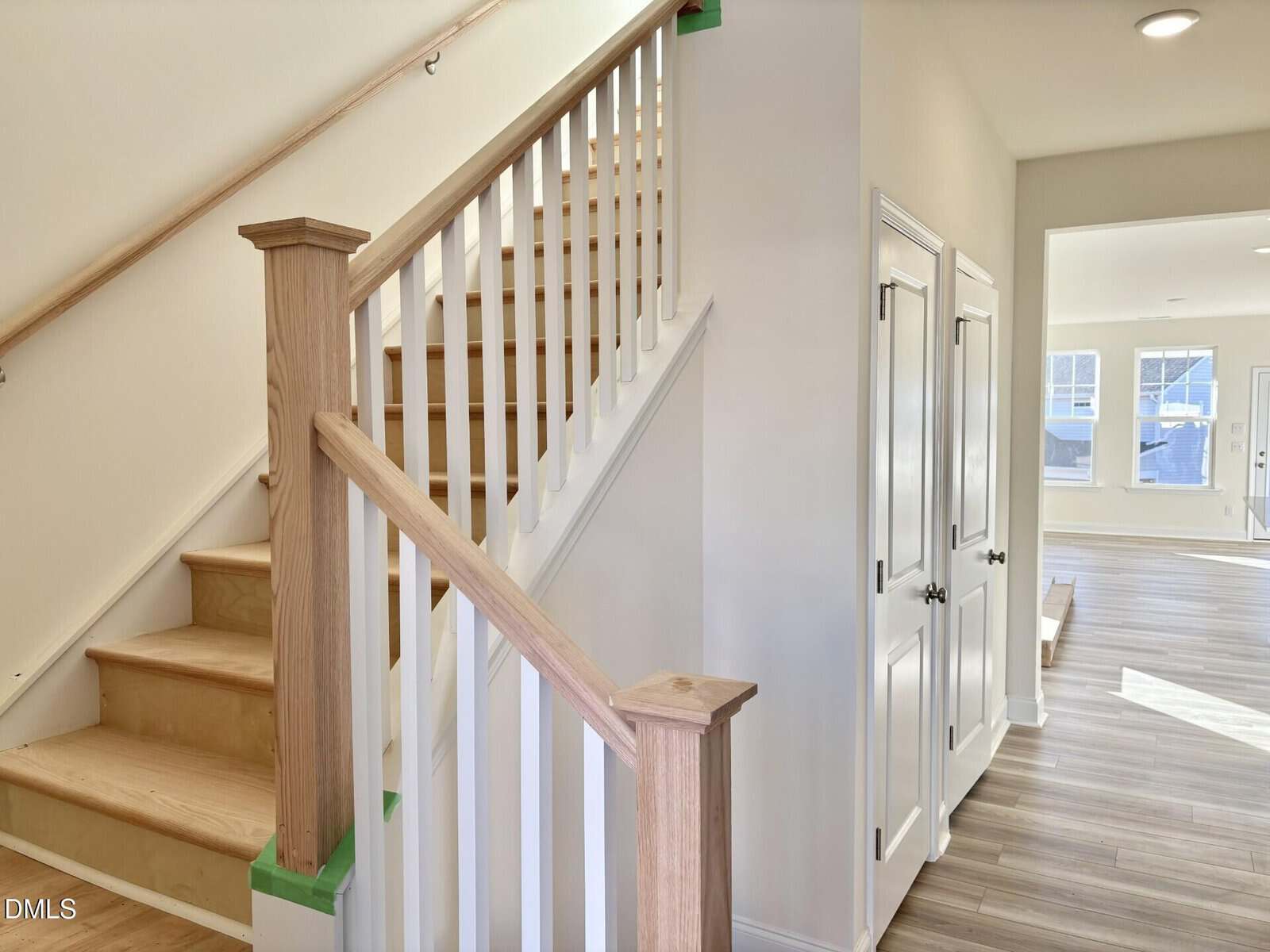 A bright, modern hallway features a wooden staircase with white railings, light-colored wood floors, double-door storage closets, and an open living area with large windows in the background.