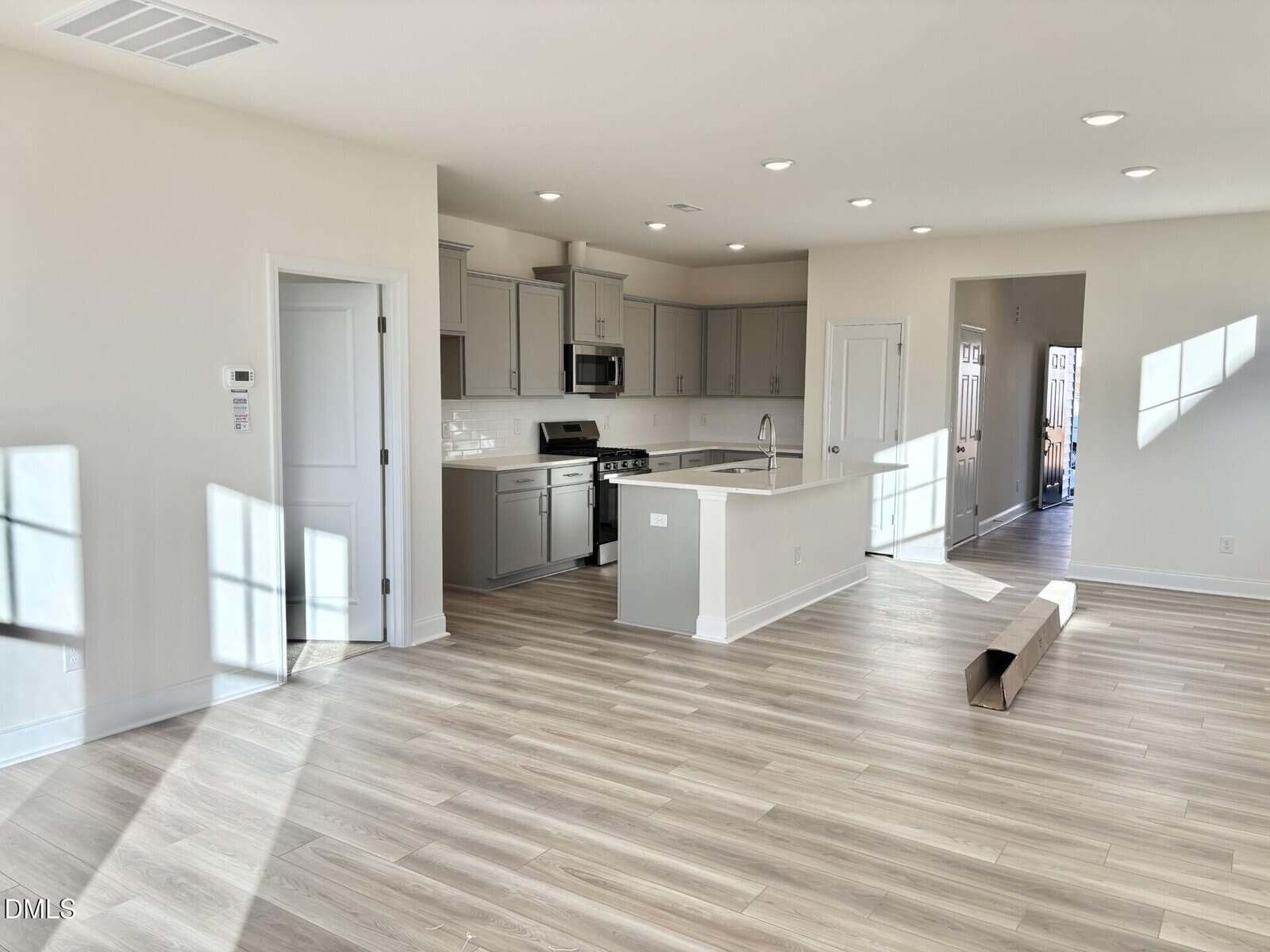 A modern, unfurnished open-plan kitchen and living space with light wood flooring, gray cabinets, stainless steel appliances, and ample natural light from several large windows. A cardboard tube lies on the floor.