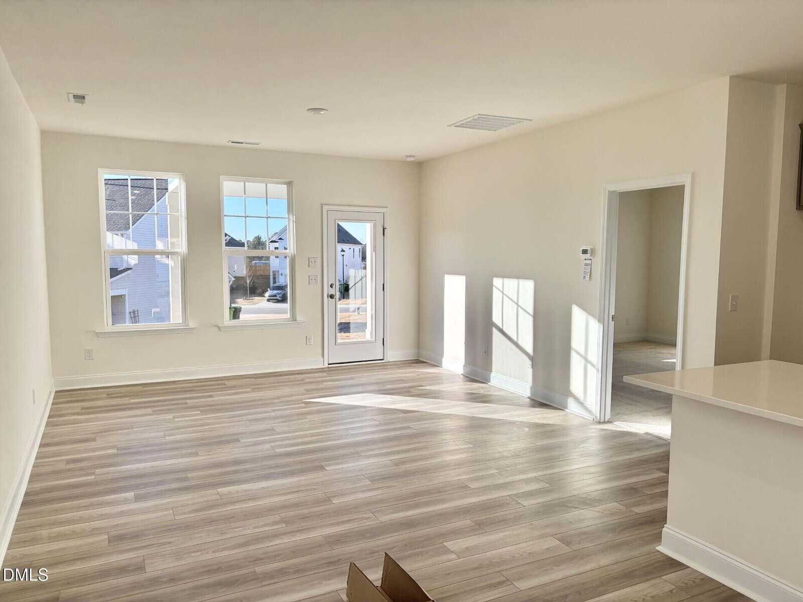 Bright, empty living room with light wood floors, large windows, white walls, and a door leading outside; kitchen countertop visible on the right, and a doorway leading to another room. Sunlight streams in through the windows.