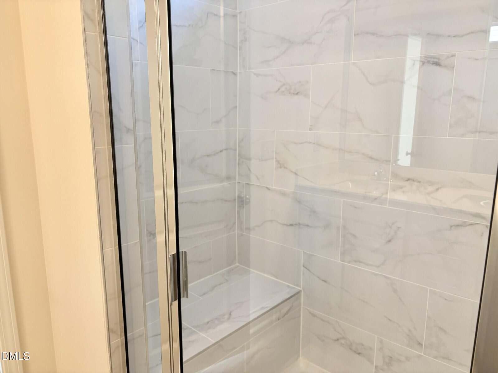 Glass shower with marble tile walls and a built-in bench. The tiles have a light gray and white veined pattern, and the shower door has a metal handle.