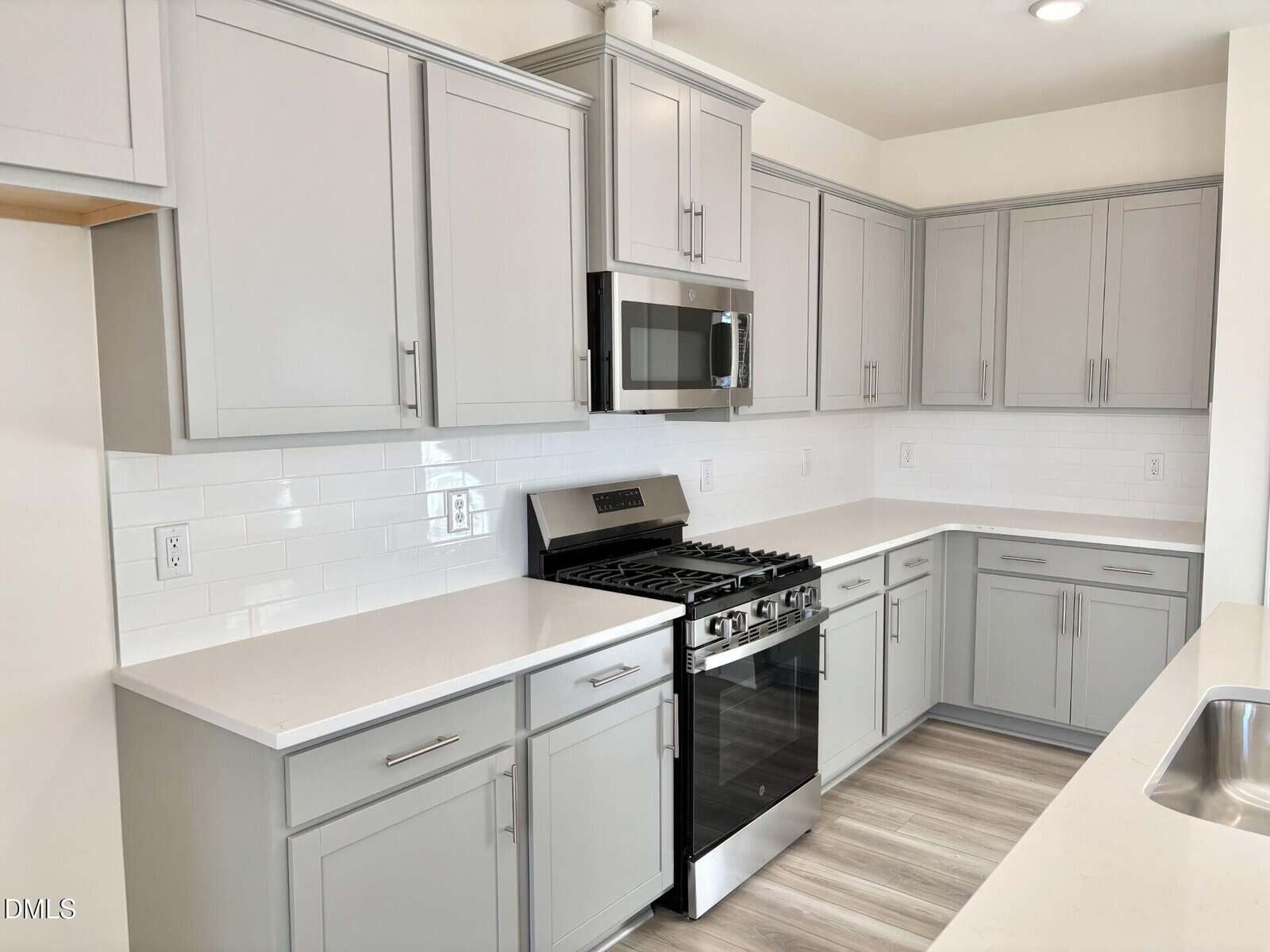 Modern kitchen with light gray cabinets, white quartz countertops, white subway tile backsplash, stainless steel gas stove and microwave, and light wood flooring. The space is clean and bright with ample cabinetry.