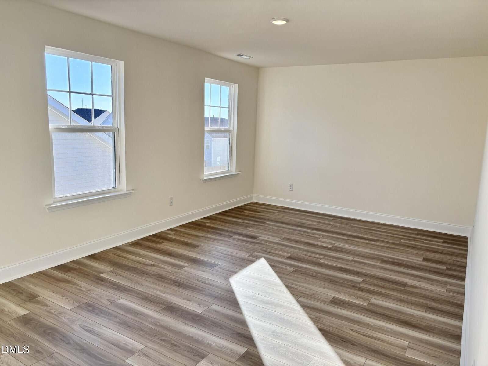 Bright, empty room with light-colored walls, two large windows, and wood laminate flooring. Sunlight streams through the windows, casting shadows on the floor. Room appears clean and modern.