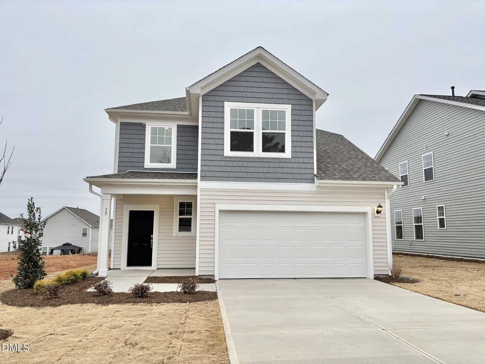 Two-story modern house with gray and white exterior, two-car garage, and large front windows, set on a plot with brown grass and minimal landscaping under a cloudy sky.