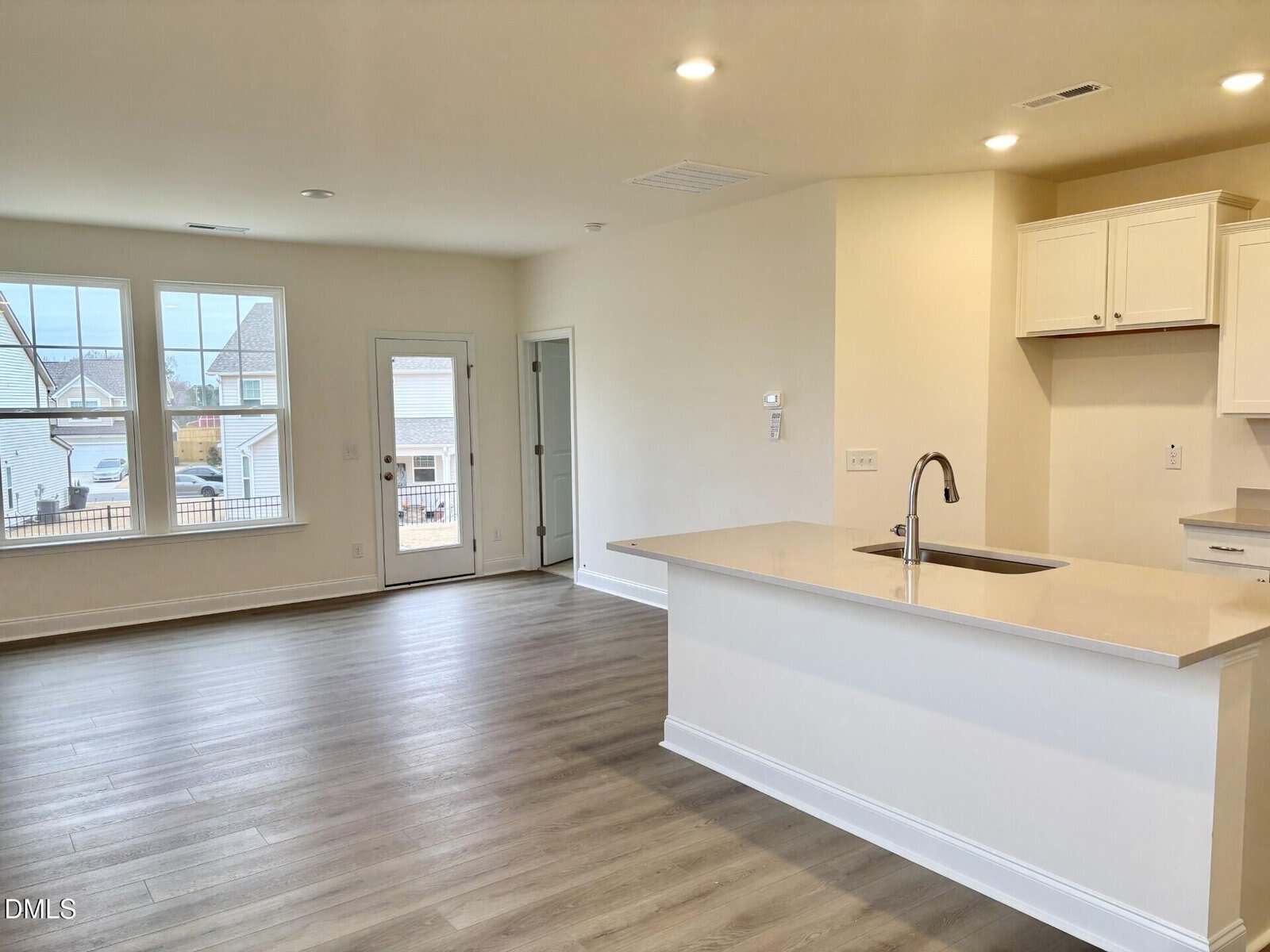 Bright, modern open-concept kitchen and living area with light wood floors, white cabinets, a large island with a sink, recessed lighting, and large windows plus a glass door leading outside.