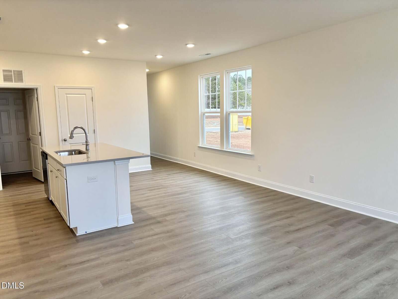 A bright, empty open-concept kitchen and living area with light wood flooring, white walls, recessed lighting, and large windows letting in natural light. A kitchen island with a sink is visible on the left.