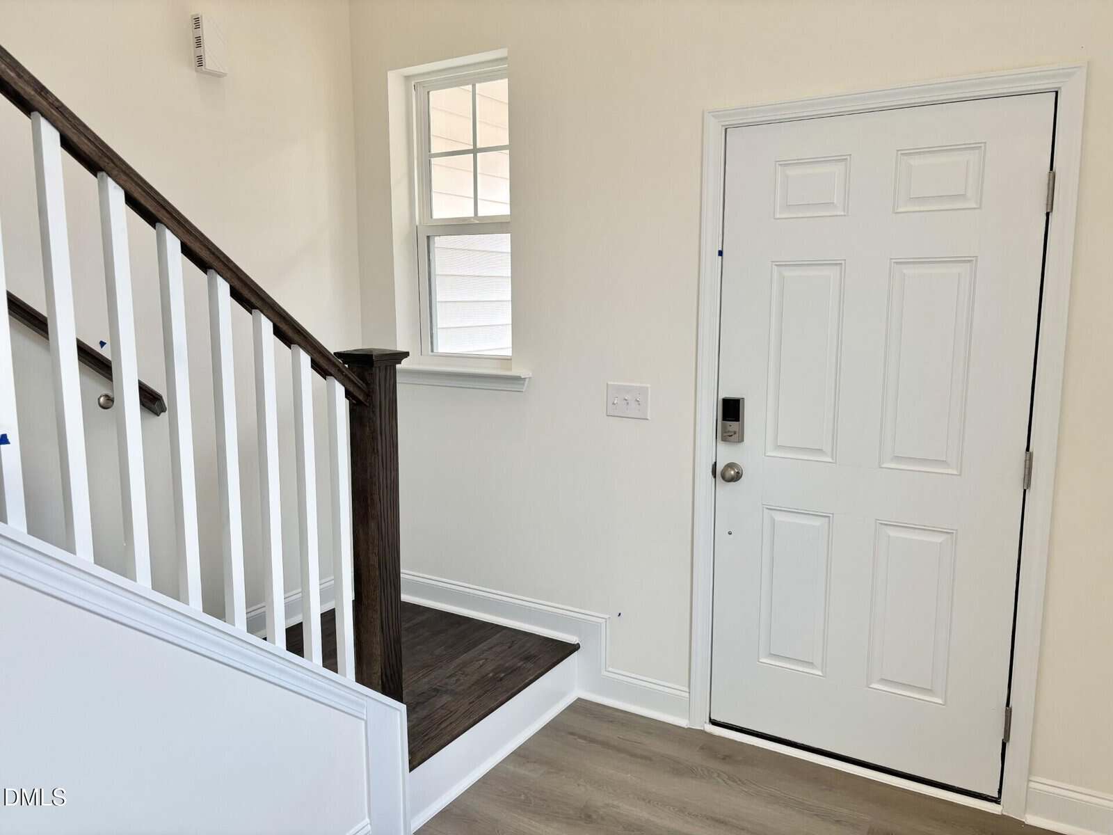 A white front door with a silver handle is beside a wooden staircase with dark handrails and white balusters. A small window is above the stairs, and the walls are painted light cream. The floor is light wood.