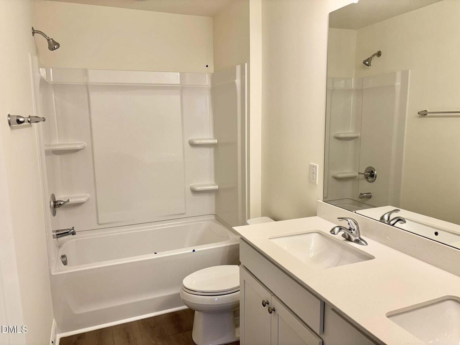 A modern bathroom with a white bathtub-shower combo, toilet, and a white vanity with a sink. There is a large mirror above the sink and wood-look flooring. The walls are light-colored.