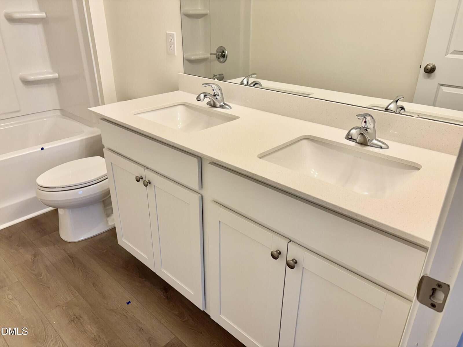 Modern bathroom with a double sink vanity, white cabinets, silver faucets, a large mirror, a toilet, and a bathtub/shower combo. The floor is wood-style, and the walls are light-colored.