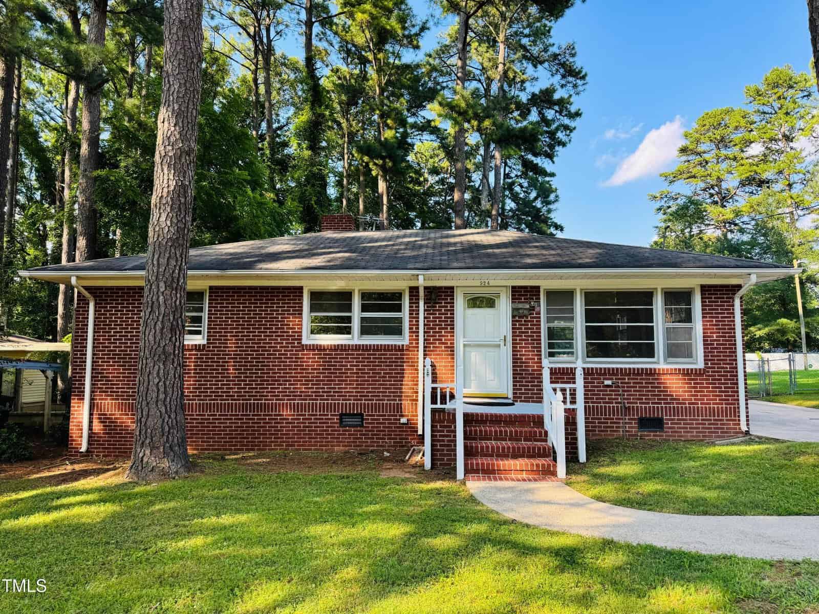 A single-story brick house at 924 Buckhorn Street in Henderson features a dark shingled roof, white-framed windows, and a central white door accessed by brick steps and railings. Tall pine trees and green grass surround the home.