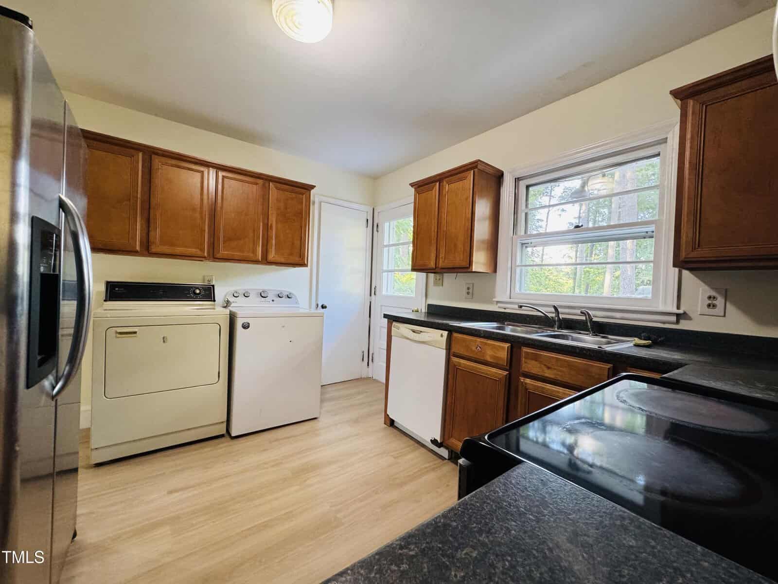 A kitchen with wood cabinets, black countertops, and light wood flooring features a washer, dryer, dishwasher, stove, and stainless steel refrigerator. A window and door are visible on the far wall.