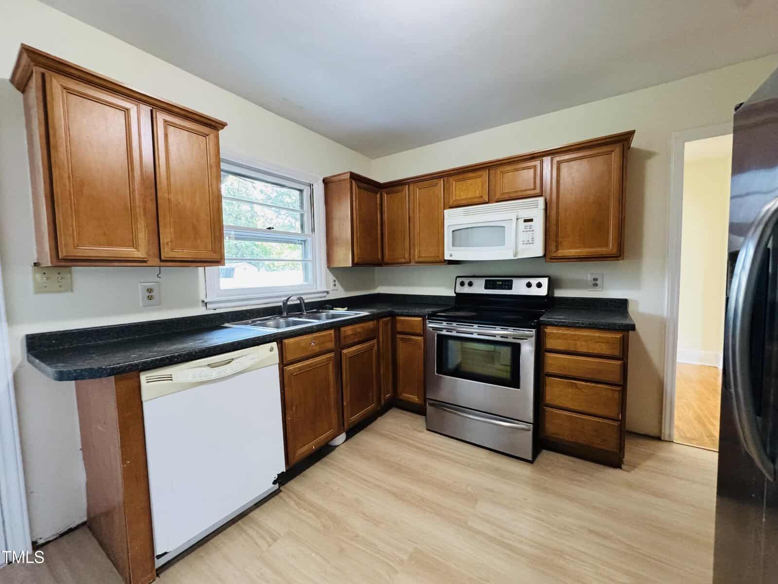 A kitchen with wood cabinets, black countertops, a stainless steel oven and microwave, a white dishwasher, a double sink under a window, and light wood flooring. Walls are painted a light color.