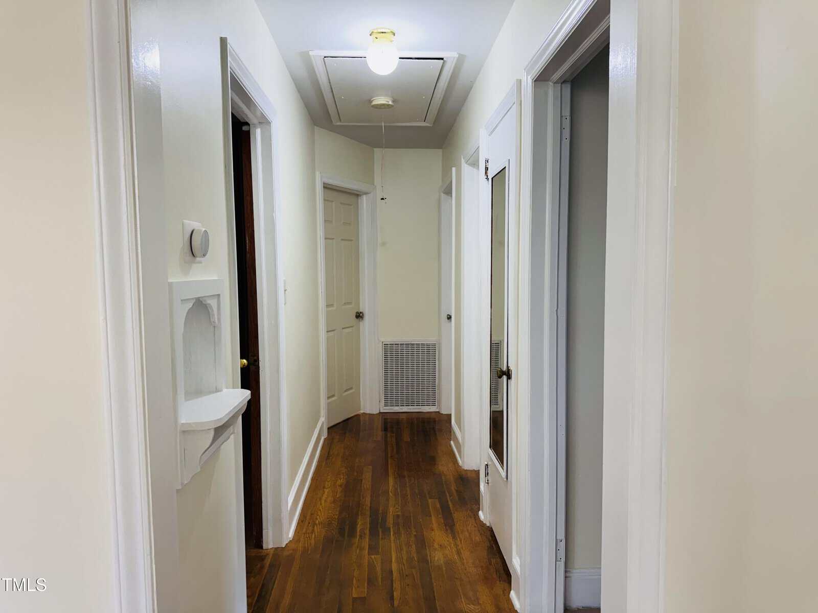 A narrow hallway with white walls and hardwood floors, several open doors on either side, a thermostat and wall niche on the left, and a ceiling light near an attic access door above.