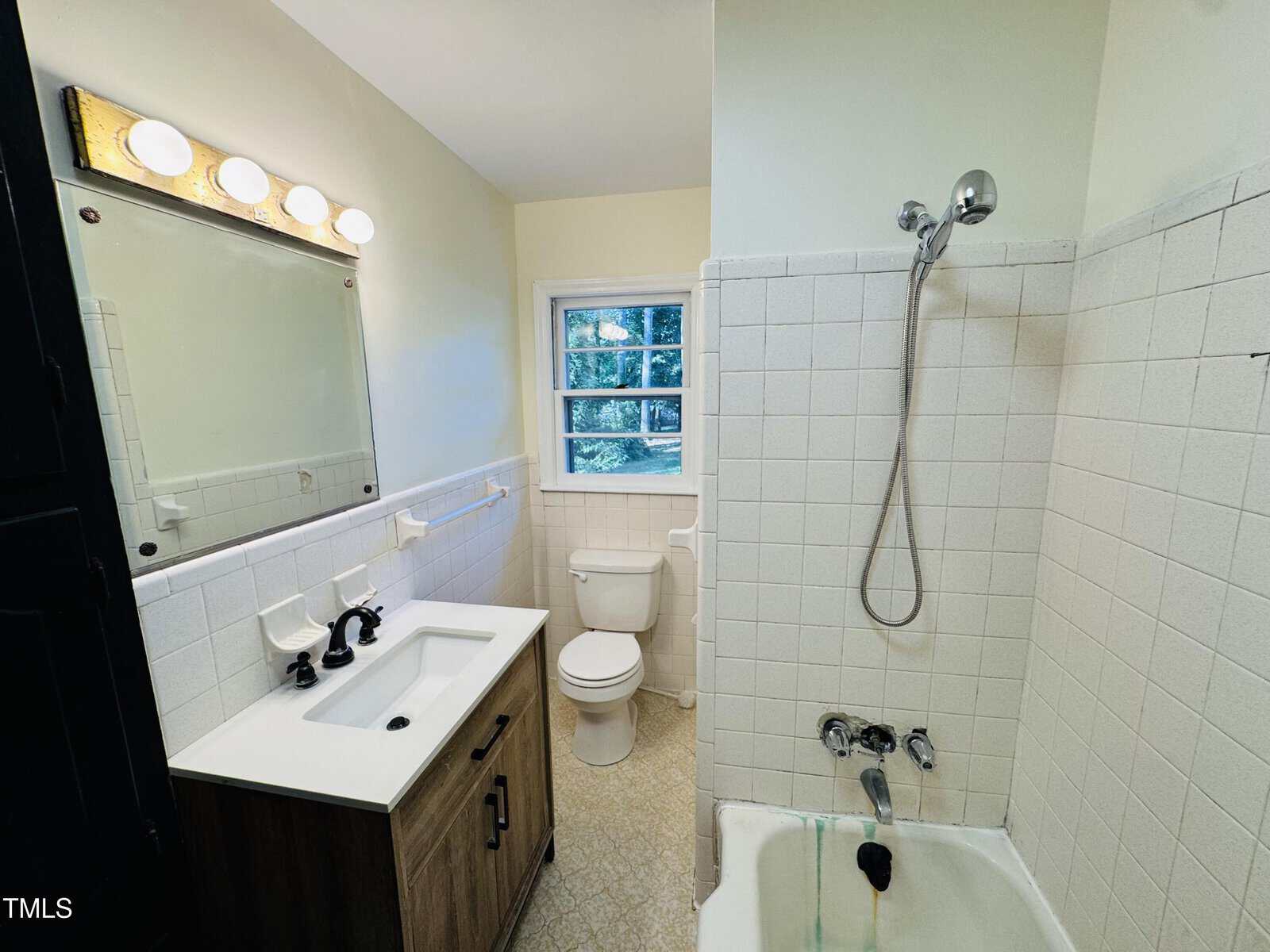 A small bathroom with a mirrored vanity, sink, toilet, and a shower-tub combo with white tile walls. There is a window above the toilet and overhead vanity lights above the mirror.